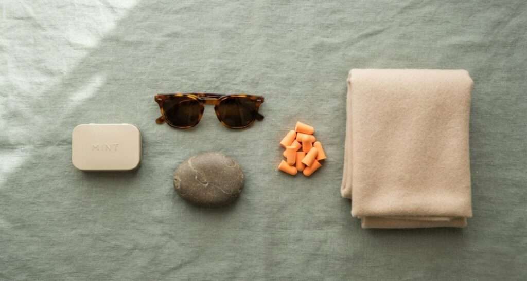 Calm flat-lay of a sensory overload reset kit — foam earplugs, sunglasses, a smooth stone, a soft scarf, and a mint tin arranged on linen.
